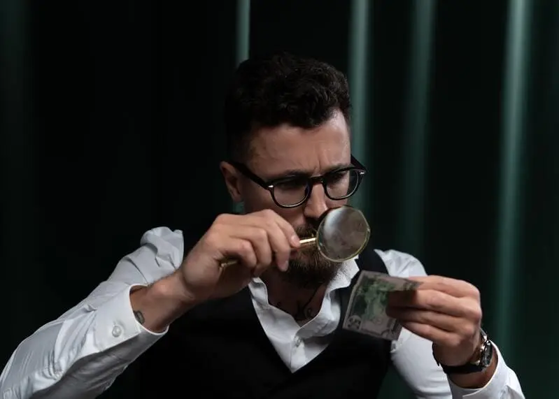 homme brun lunettes barbe tenant loupe et billet de banque